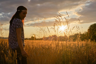 Young woman standing on wheat field against cloudy sky during sunset