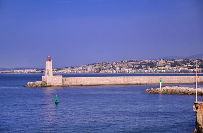 Lighthouse by sea against buildings against clear blue sky