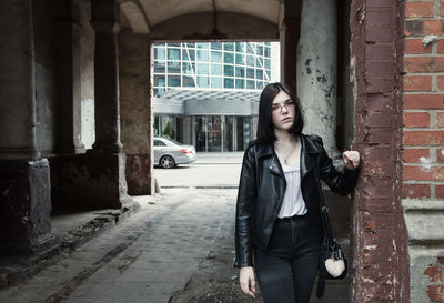 Sad young girl in black jacket and jeans stands in old arch on city street on summer day