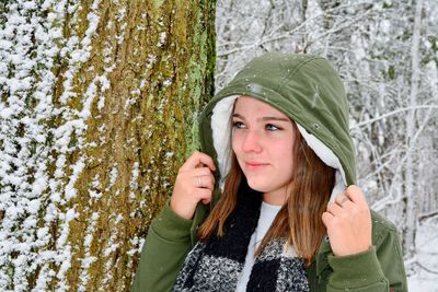 Young woman in hood standing by tree trunk during winter