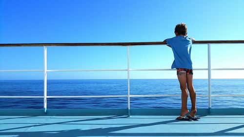 Rear view of boy standing by railing at boat in sea against clear blue sky