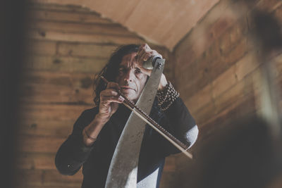 Low angle portrait of mature man playing music instrument against wooden wall