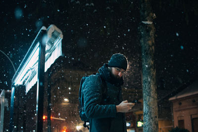 Man using mobile phone while standing against building during snowfall at night