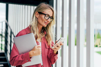 Confident businesswoman uses phone while standing with laptop at exit from office building.