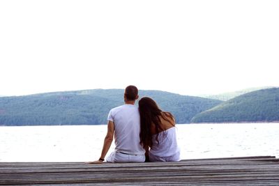 Young couple sitting on pier while looking at lake against sky