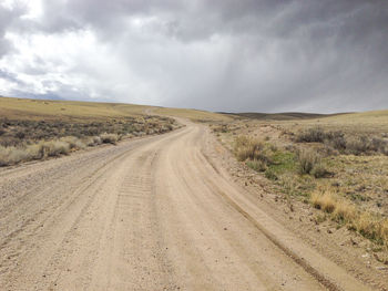 Empty dirt road amidst field against sky