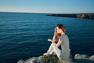 Woman sitting on rock by sea against clear sky