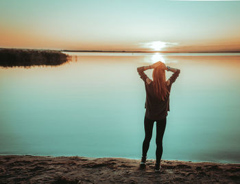 Rear view of woman standing at lakeshore against sky during sunset