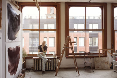 Thoughtful artist holding coffee cup while sitting by window on table at workshop