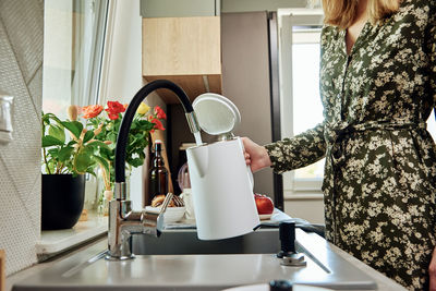 Woman pouring water from faucet into electric kettle at the kitchen