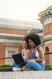 Happy friends using laptop while sitting on retaining wall