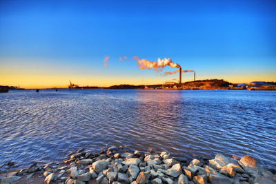 Smoke stack by sea against sky during sunset