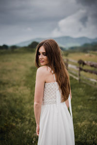 Side view of young woman standing on grassy field against cloudy sky