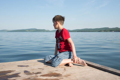 The boy sits on a wooden pier and looks at the water of the lake on a sunny day.