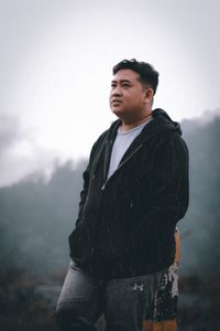 Young man looking away while standing in rain against sky