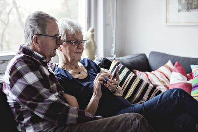 Retired senior couple sharing smart phone sitting on sofa in living room at home