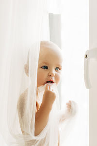 A little boy is standing on the windowsill near the window, hiding behind the curtain