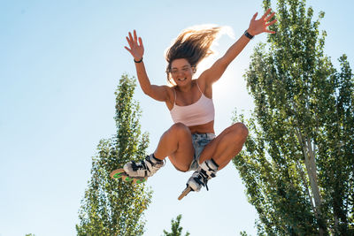 From below of active female in rollerblades jumping and performing trick in park against blue sky in summer on sunny day