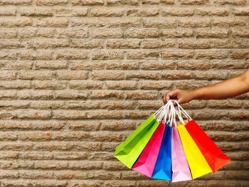 Cropped hand of woman holding colorful shopping bags against stone wall