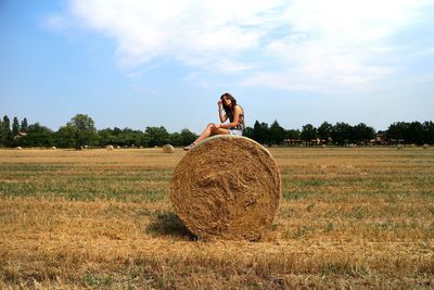 Woman sitting on hay bale at field against sky