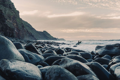 Figure of a female surfer walking on a cobble's beach at sunset