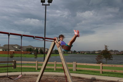 Girl enjoying swing at playground by lake against cloudy sky