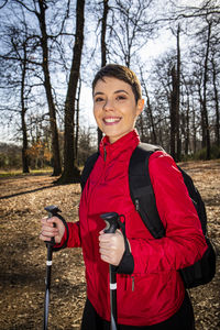 Young woman has short hair, wears a red jacket and uses trekking poles for a hike in the woods