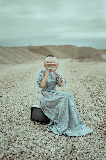 Portrait of woman holding fish bowl while sitting on field against cloudy sky