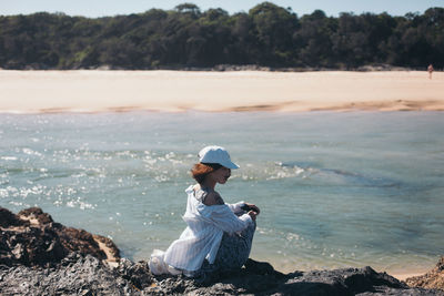 Side view of woman sitting on rock at beach during summer