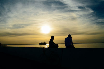 Silhouette men sitting on shore against sky during sunset