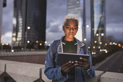 Woman with tablet pc standing on bridge in city at dusk