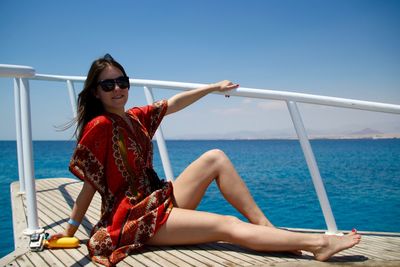 Full length portrait of beautiful woman sitting on pier by sea against clear blue sky