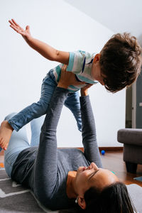 Happy woman laughing and lifting adorable boy while lying on floor in cozy room at home