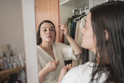 Hispanic woman in white bathrobe applying makeup to her eyelids with small brush in front of mirror 