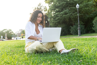 Happy young hispanic woman in stylish clothes smiling and browsing data on laptop while sitting on grass on summer day in park