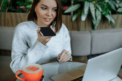 A young woman is working on a laptop in a cafe. records an audio message on a smartphone