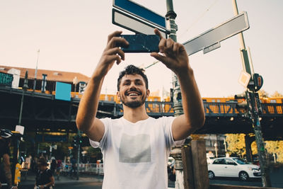 Smiling young man photographing through mobile phone while standing on street in city