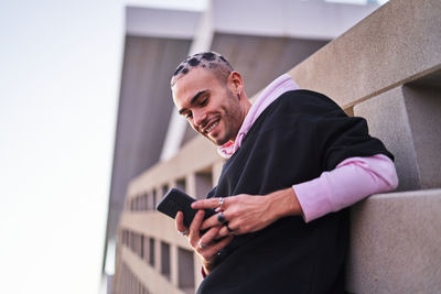 Low angle of trendy young male in hoodie using mobile phone and leaning on concrete wall and smiling widely