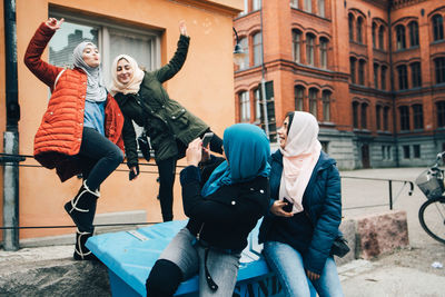 Young woman photographing cheerful female friends against building in city