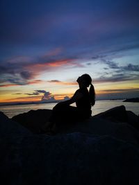 Silhouette man sitting on rock by sea against sky during sunset