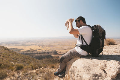 Side view of young man shielding eyes with hat while sitting on mountain