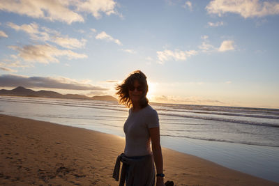 Rear view of woman standing at beach against sky during sunset