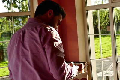 Rear view of man using mobile phone while standing against window