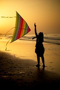 Silhouette girl holding kite while standing at beach against sky during sunset