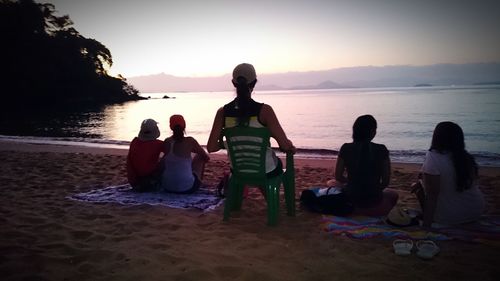 Rear view of people sitting on beach against sky during sunset