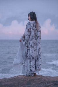 Rear view of woman standing at beach against sky during sunset