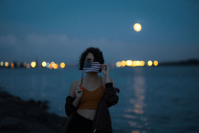 Portrait of woman holding flag while standing at beach against sky at dusk