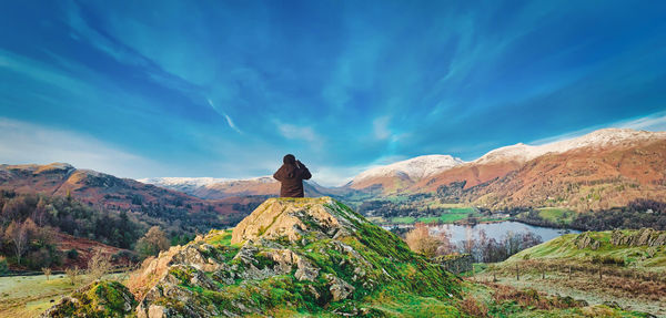 Rear view of man sitting on rock looking at mountain range against sky