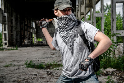 Young man wearig mask throwing bottle while standing under bridge