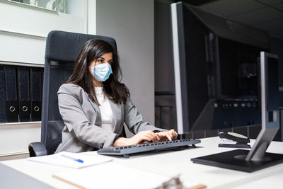 Confident young female specialist in formal suit and protective mask sitting at desk and using computer while working in modern workspace